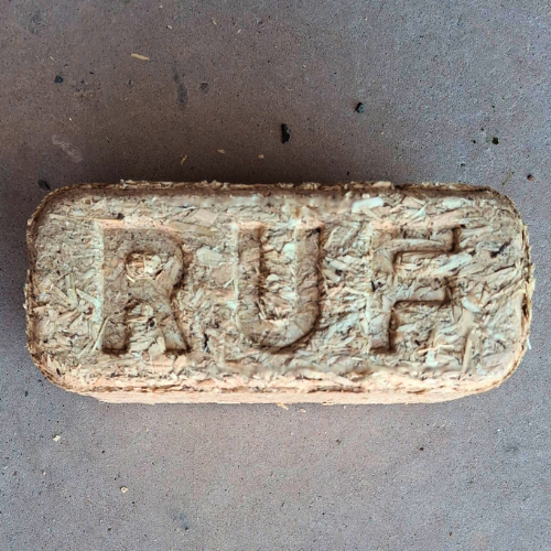 Firewood briquette in the shape of a rectangular block with the "RUF" brand stamped thereon. The briquette texture is of compressed wood chips which has been placed on a grey concrete background. 