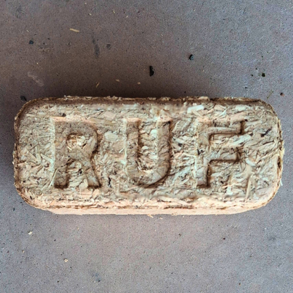 Firewood briquette in the shape of a rectangular block with the "RUF" brand stamped thereon. The briquette texture is of compressed wood chips which has been placed on a grey concrete background. 